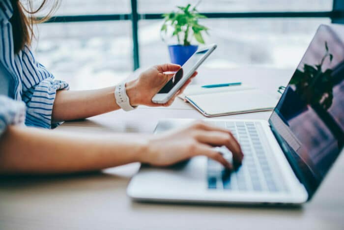 Hands of a woman, holding a cell phone and typing on a laptop